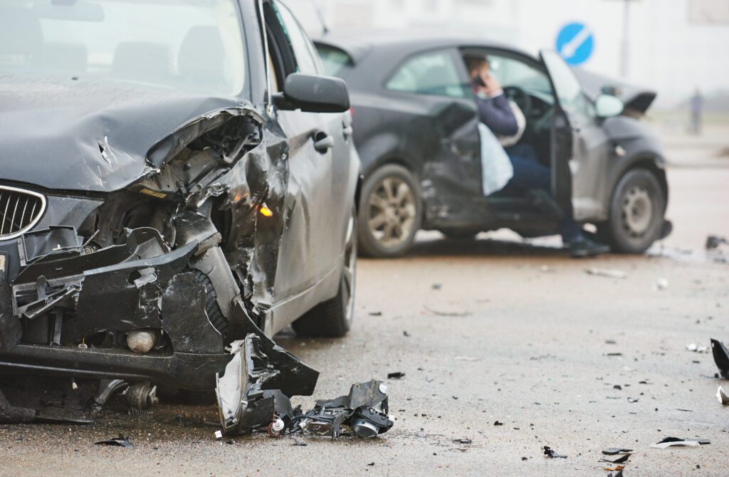 Two damaged cars after an accident with debris scattered and a person standing in background.