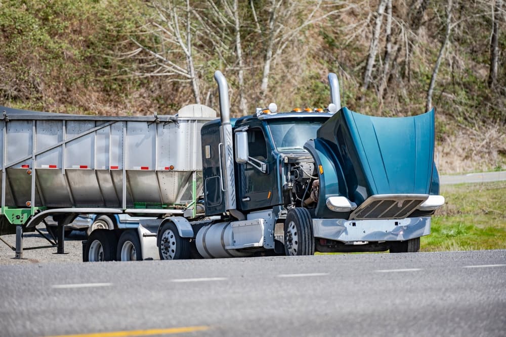 An industrial dark green big rig with a day cab and a loaded bulk semi trailer is broken down, standing out of service on the road shoulder with its hood open, waiting for assistance.






