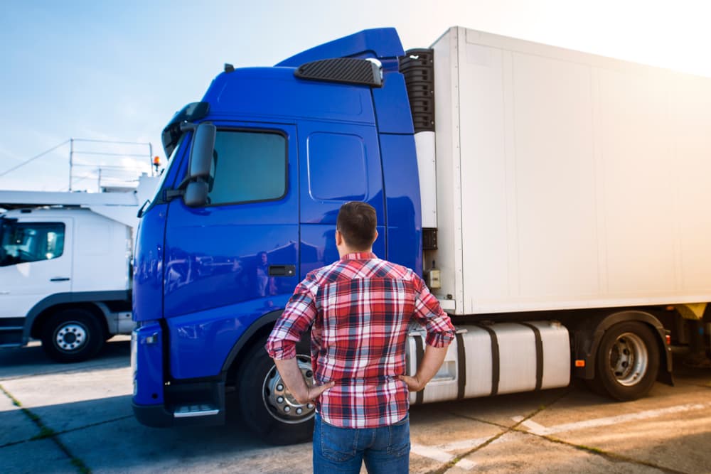 A professional middle-aged truck driver in casual clothes is looking at his truck vehicle, preparing for a long transportation drive.