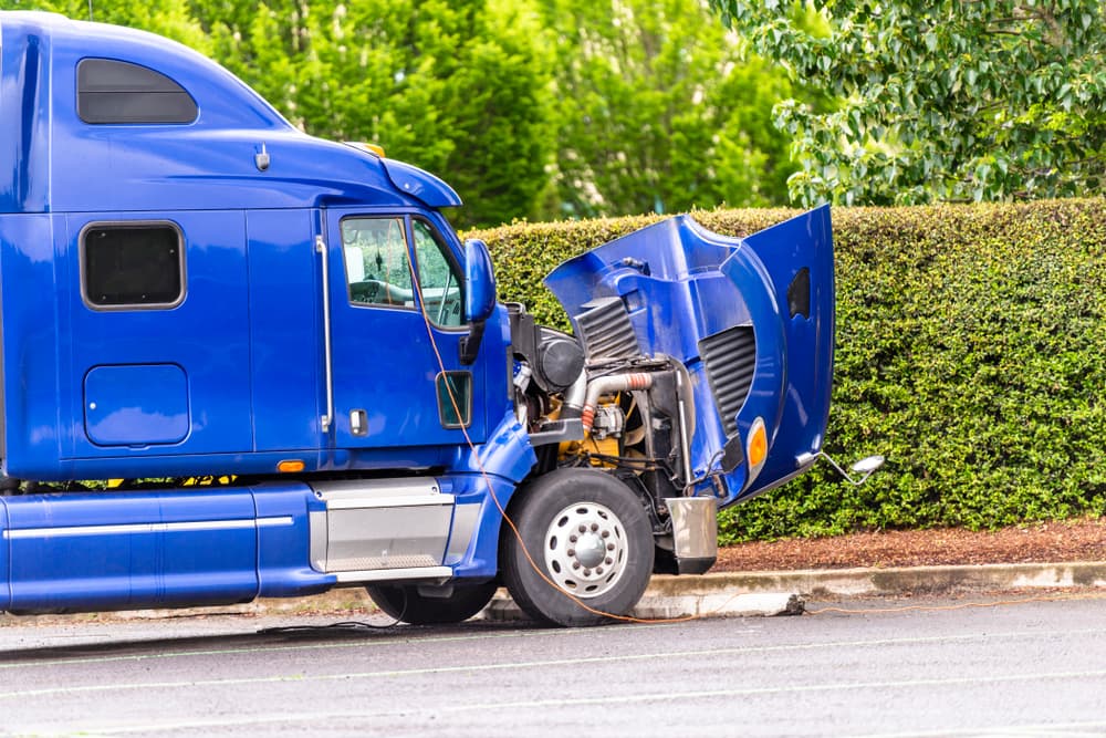 A broken American industrial-grade freight transportation blue big rig semi truck tractor with an open hood stands in the truck stop parking lot.